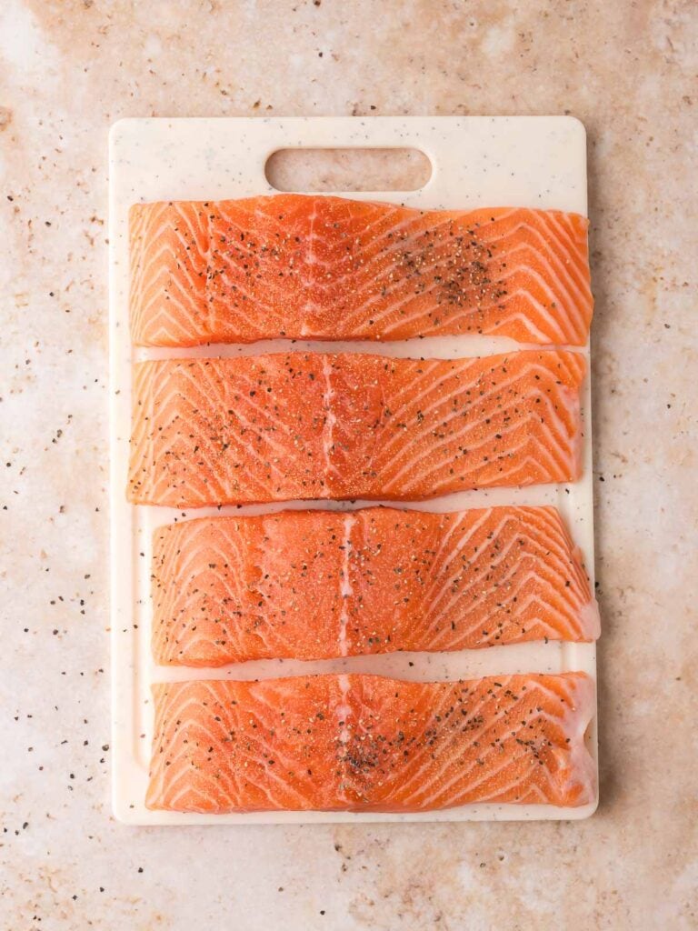Four raw salmon fillets seasoned with salt, pepper, and garlic powder on a white cutting board before cooking for Air Fryer Sweet Chili Salmon.