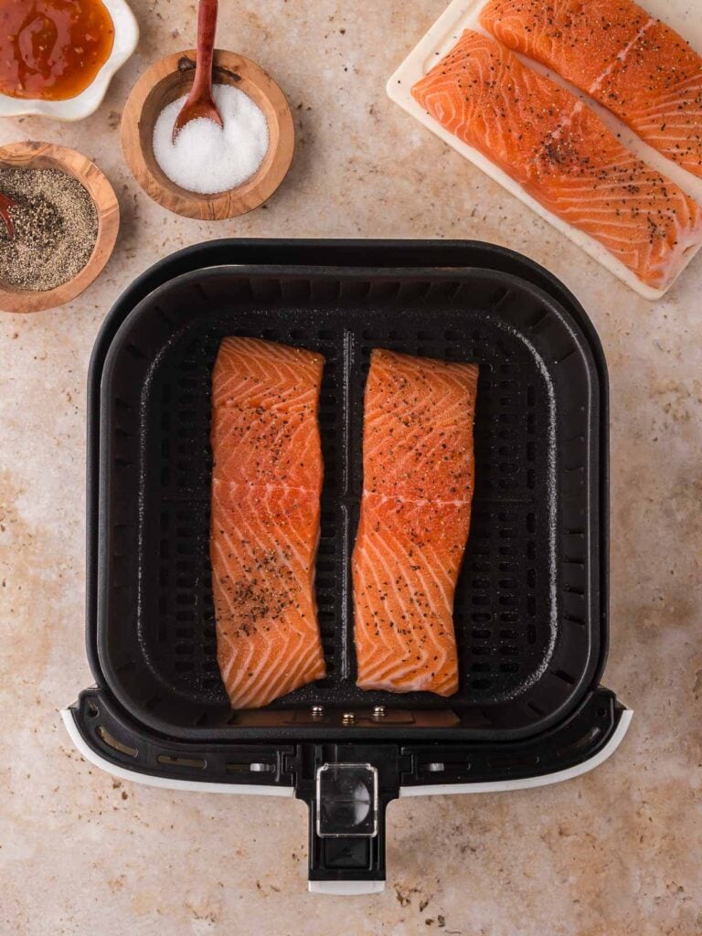 Two seasoned salmon fillets sitting in an air fryer basket with sweet chili sauce, salt, and pepper laid out beside them for Air Fryer Sweet Chili Salmon.