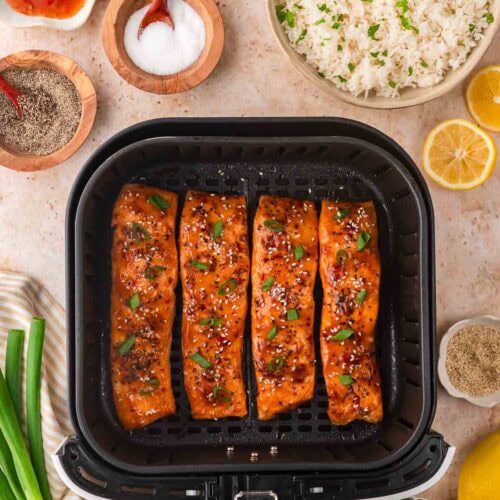 Four sweet chili glazed salmon fillets cooked in the air fryer and topped with sesame seeds and green onions, surrounded by rice, lemon, and seasoning ingredients for Air Fryer Sweet Chili Salmon.