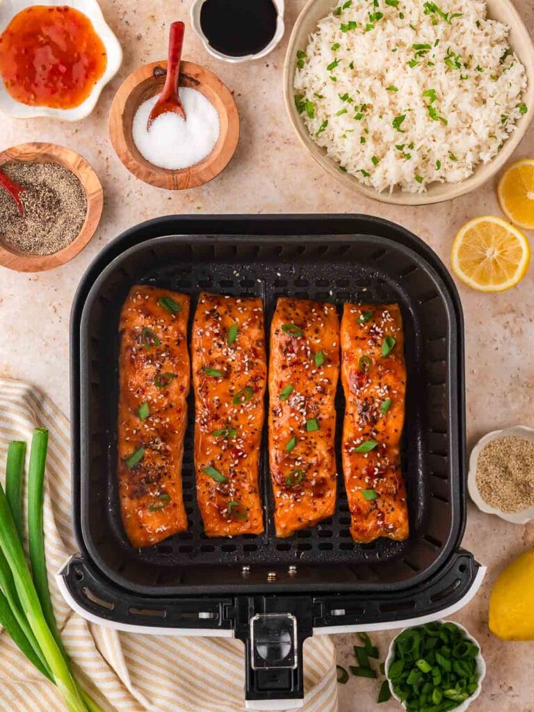 Four sweet chili glazed salmon fillets cooked in the air fryer and topped with sesame seeds and green onions, surrounded by rice, lemon, and seasoning ingredients for Air Fryer Sweet Chili Salmon.