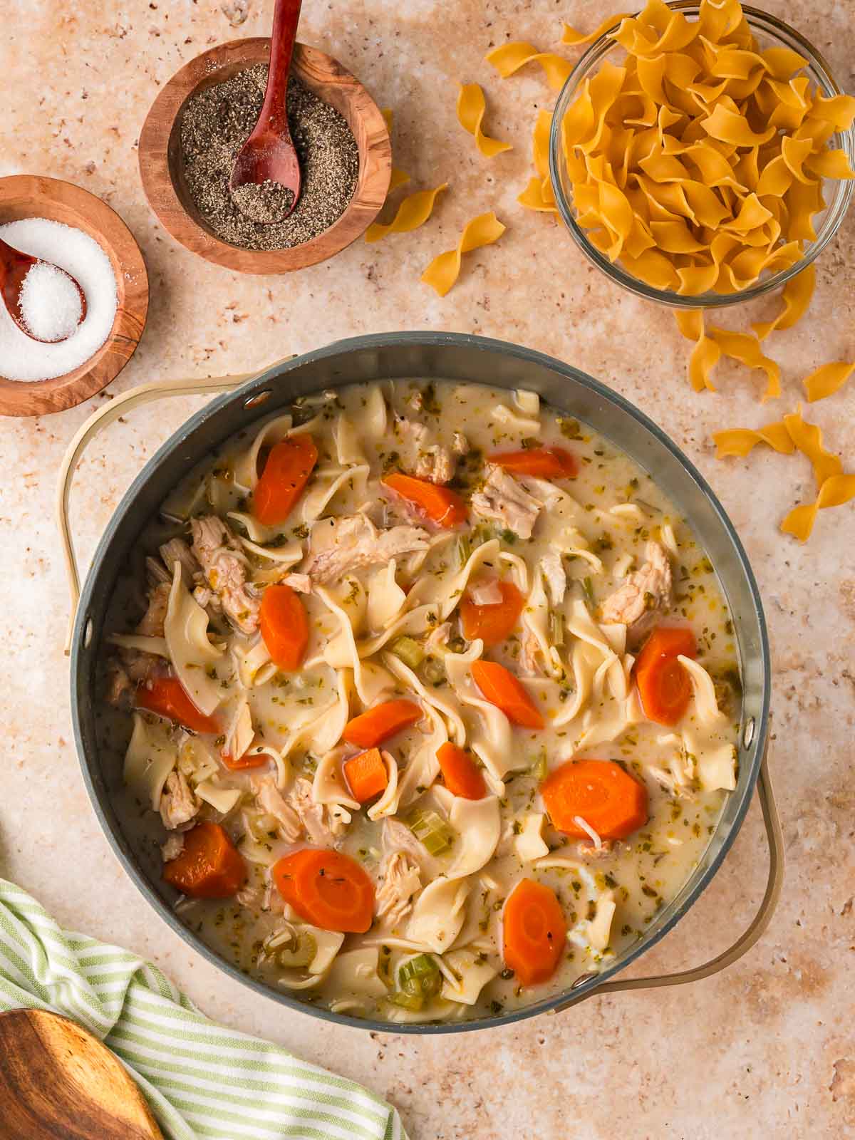 Creamy chicken noodle soup in a large pot with the creamy mixture just added, surrounded by egg noodles, salt, and pepper on a kitchen counter.