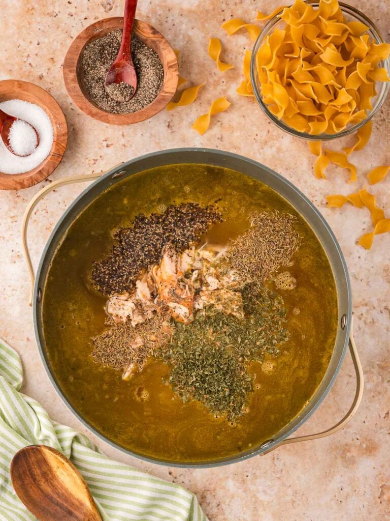 Shredded chicken, dried herbs, and seasoning added to a pot of hot broth while making high protein chicken noodle soup.