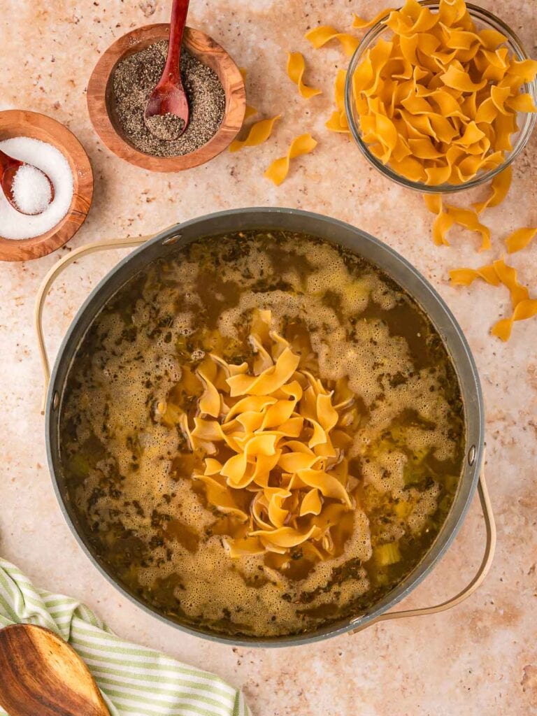 Egg noodles being added to a simmering pot of chicken broth with vegetables and herbs for creamy chicken noodle soup.