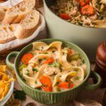Creamy chicken noodle soup served in a green bowl with a pot of soup and slices of bread in the background.
