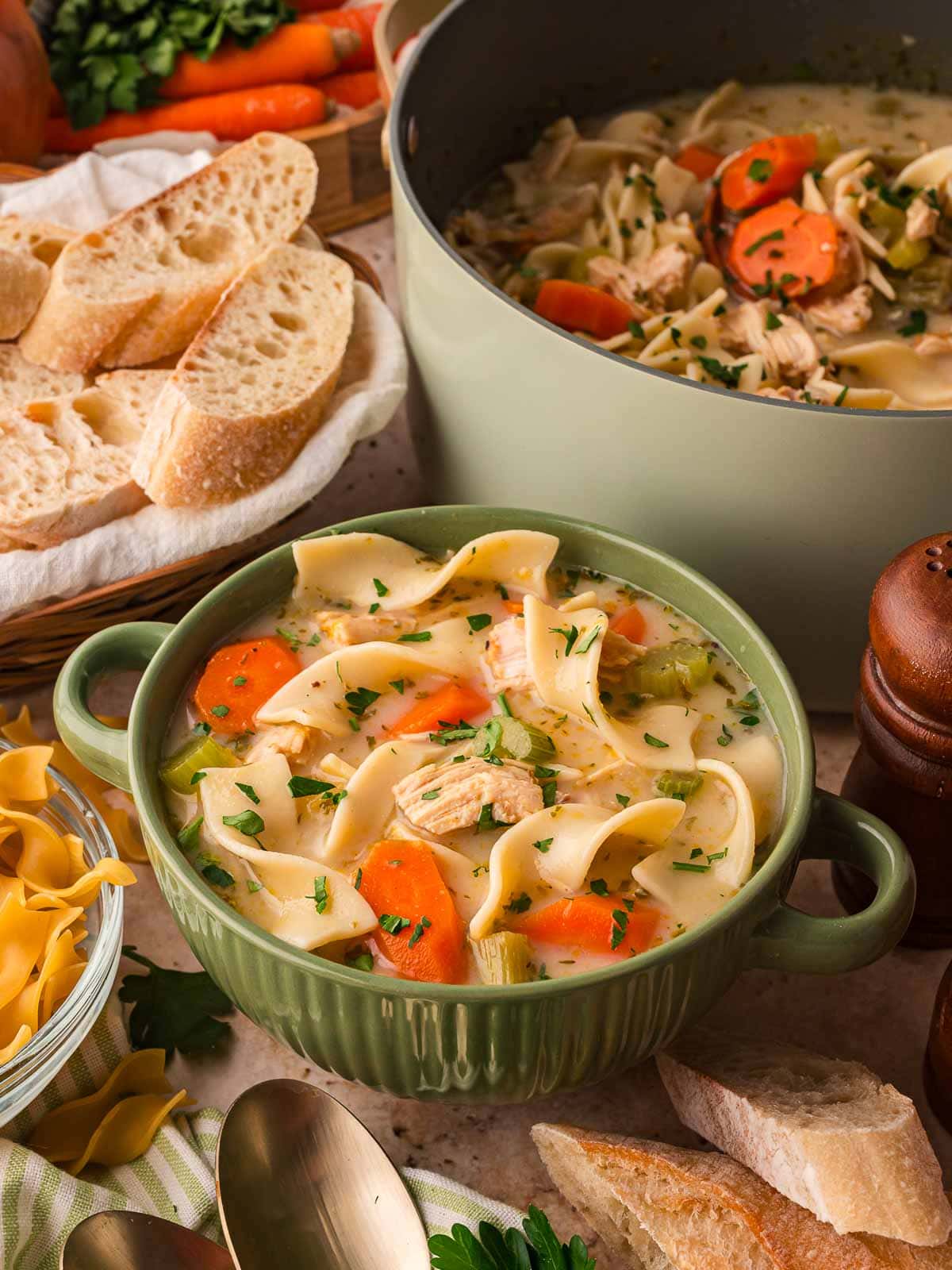 Creamy chicken noodle soup served in a green bowl with a pot of soup and slices of bread in the background.
