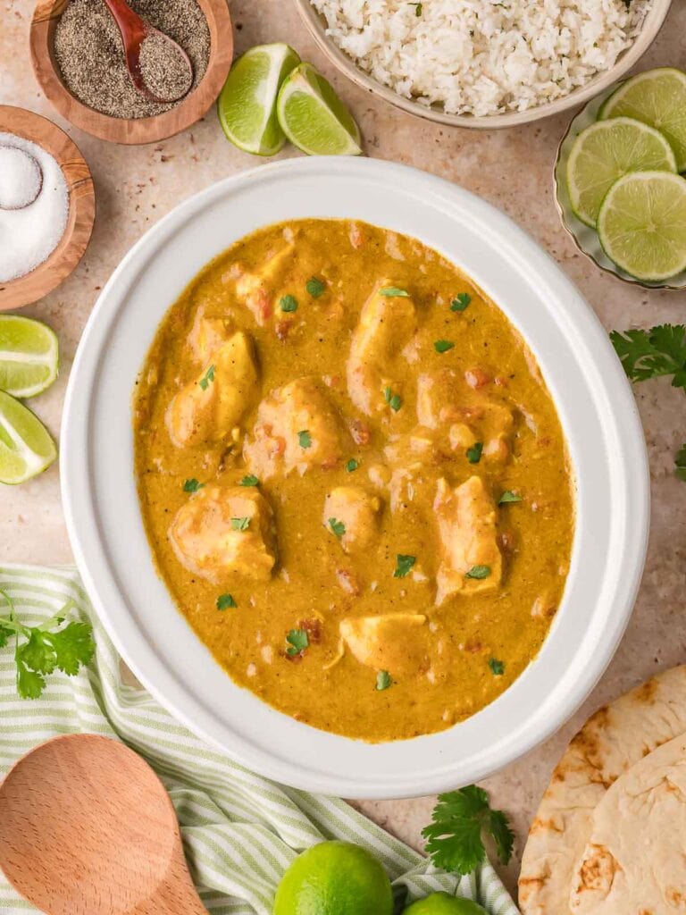 Crockpot filled with coconut chicken curry surrounded by bowls of rice, lime wedges, and fresh cilantro.