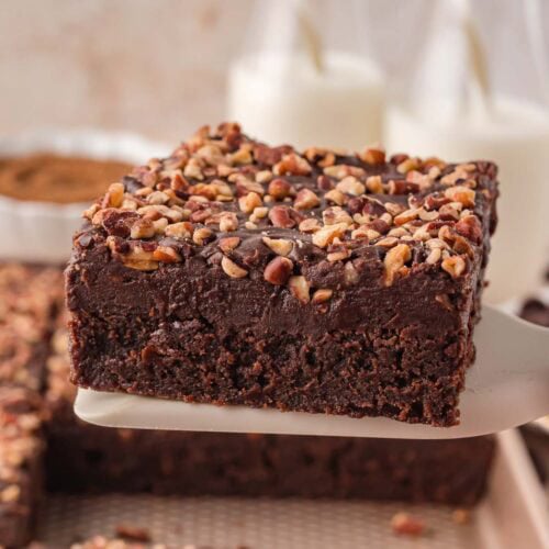 Close up of a Texas Sheet Cake slice on a spatula with a smooth chocolate frosting layer and chopped pecans, with milk bottles in the background.