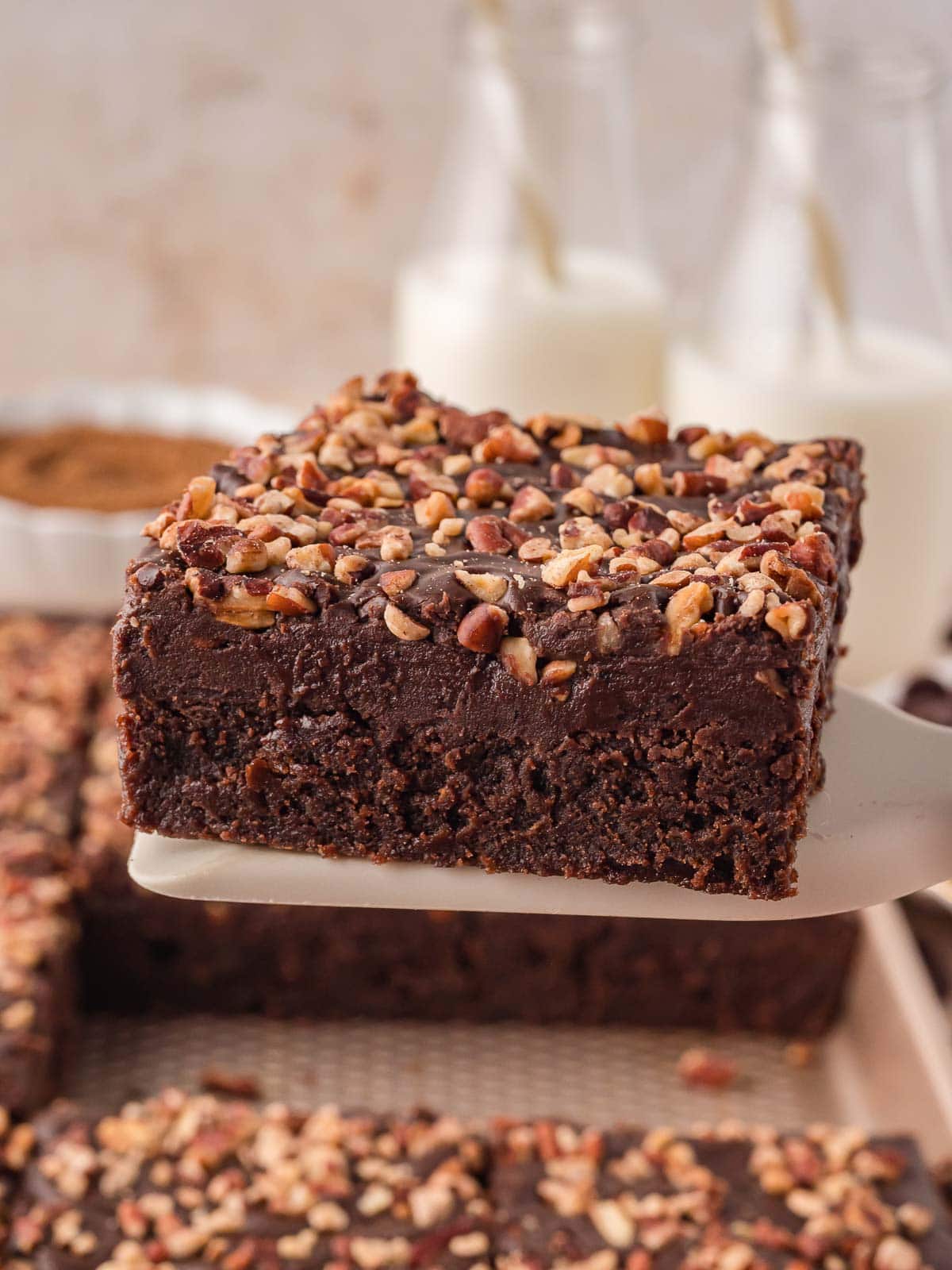 Close up of a Texas Sheet Cake slice on a spatula with a smooth chocolate frosting layer and chopped pecans, with milk bottles in the background.