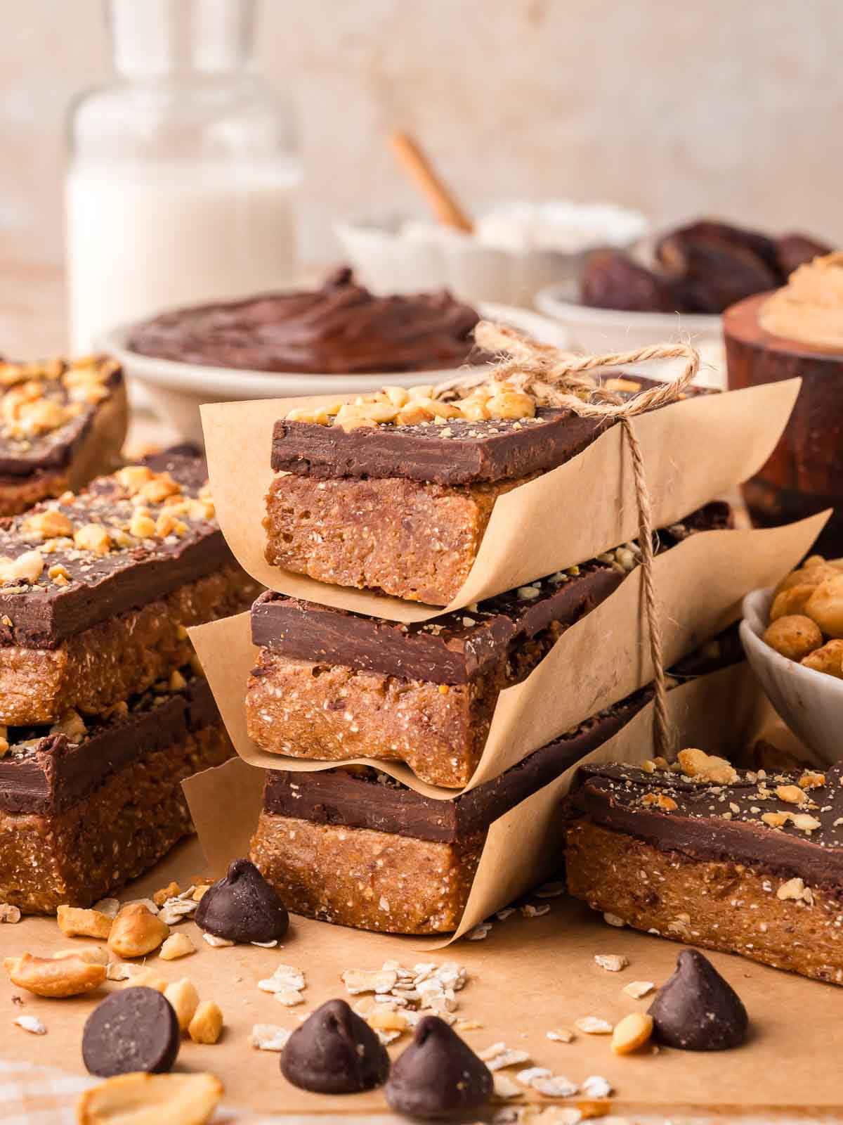 Chocolate peanut butter protein bars wrapped with parchment and twine, surrounded by peanuts, chocolate chips, and oats.