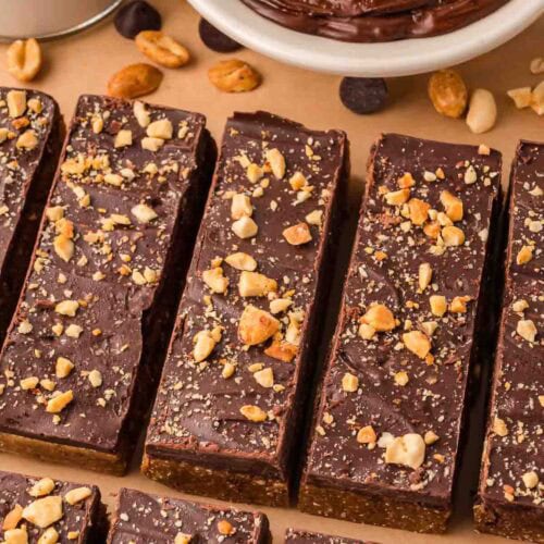 Chocolate peanut butter protein bars topped with crushed peanuts and a thick chocolate layer on a wooden board.