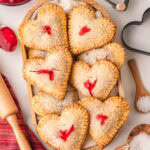Heart shaped cherry hand pies on a wooden tray with rolling pin, cherry pie filling, and heart shaped cookie cutters.