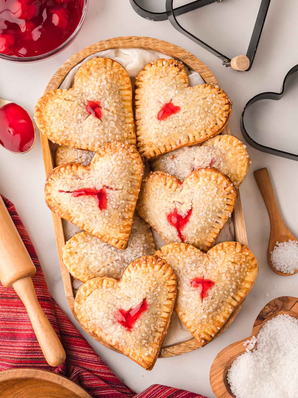 Heart shaped cherry hand pies on a wooden tray with rolling pin, cherry pie filling, and heart shaped cookie cutters.