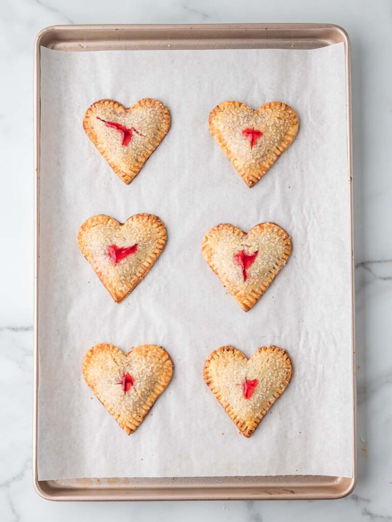 Baked heart-shaped cherry hand pies with golden crusts cooling on a parchment-lined baking sheet.