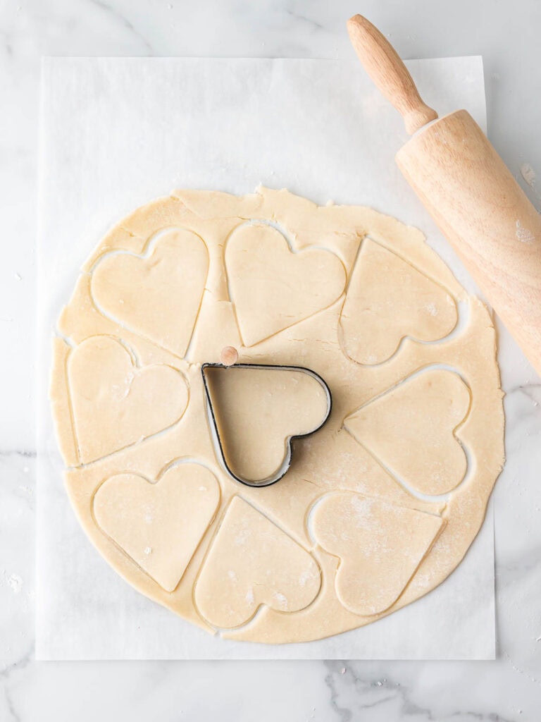 Rolled-out pie dough on parchment paper with heart shapes cut out using a heart-shaped cookie cutter.