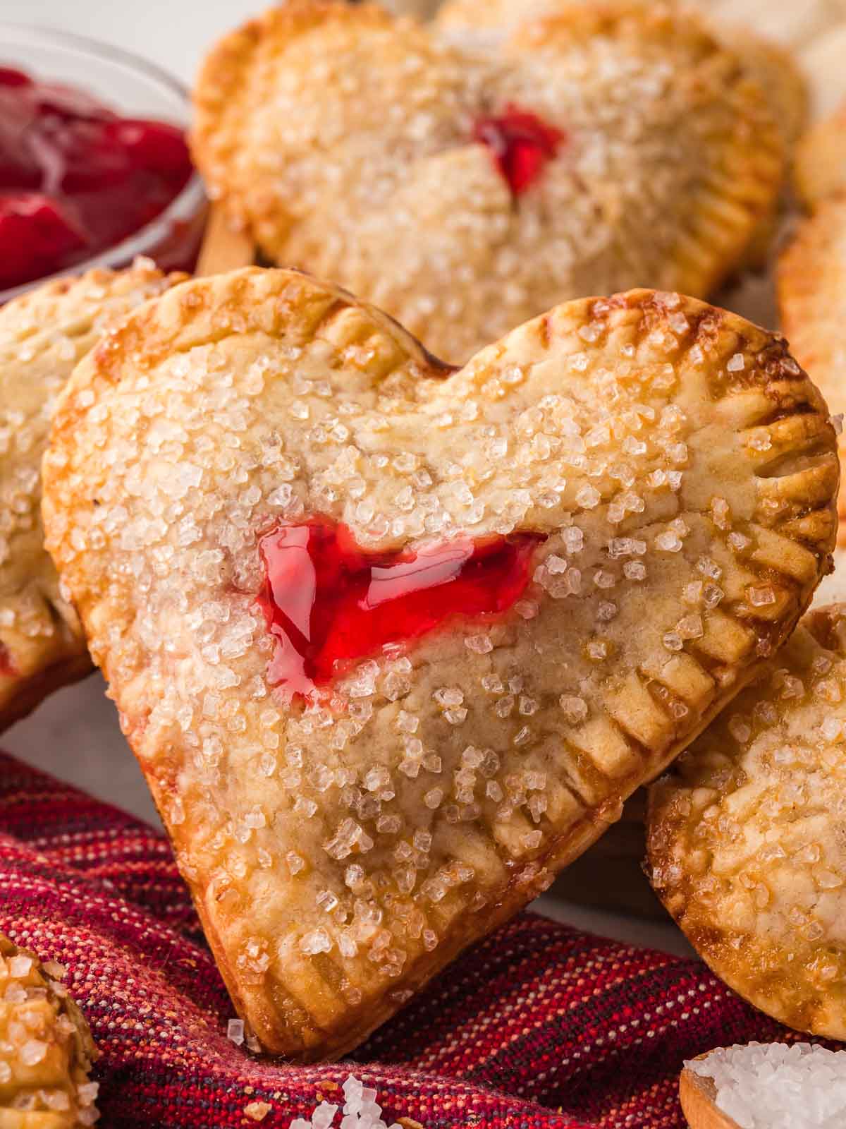 Heart shaped cherry hand pies baked until golden and topped with coarse sugar, shown stacked on a plate.