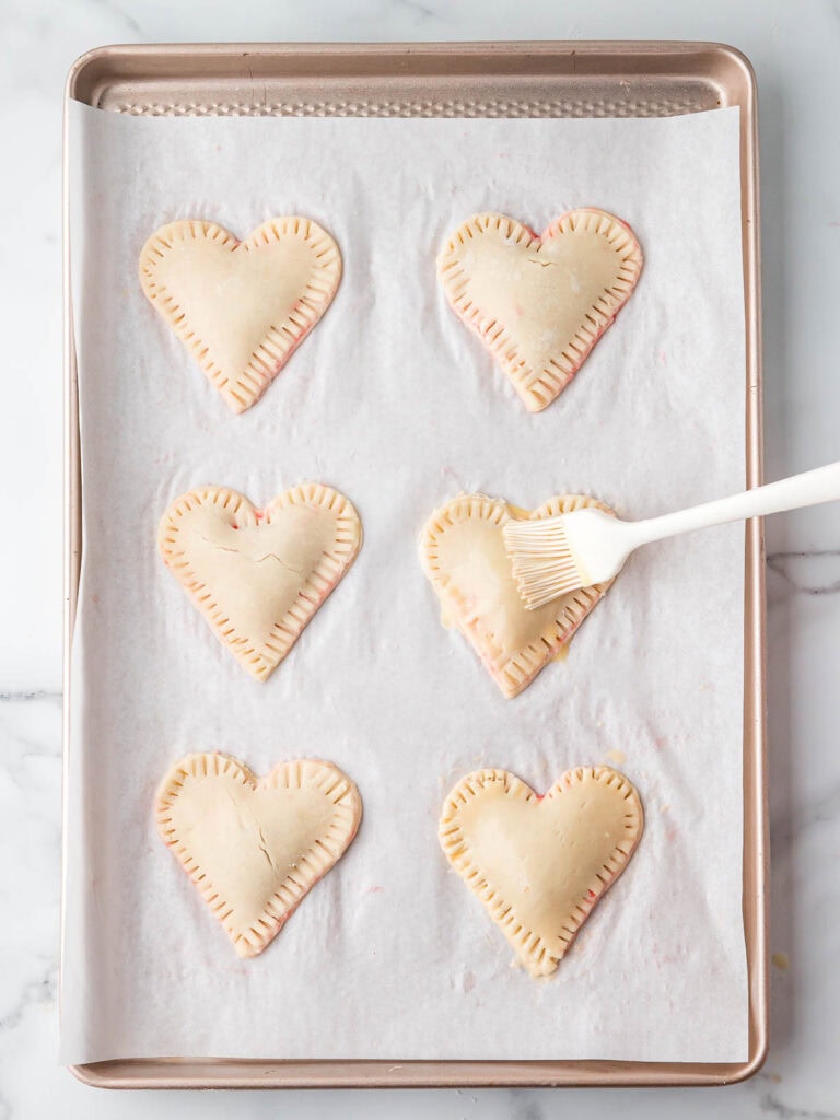 Egg wash being brushed onto heart-shaped cherry hand pies before baking.