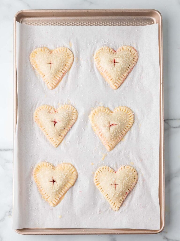 Unbaked heart-shaped cherry hand pies sprinkled with coarse sugar and vented in the center on a baking sheet.
