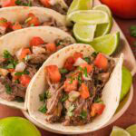 Plate of shredded beef tacos with diced tomatoes, onion, and cilantro, served with lime wedges.