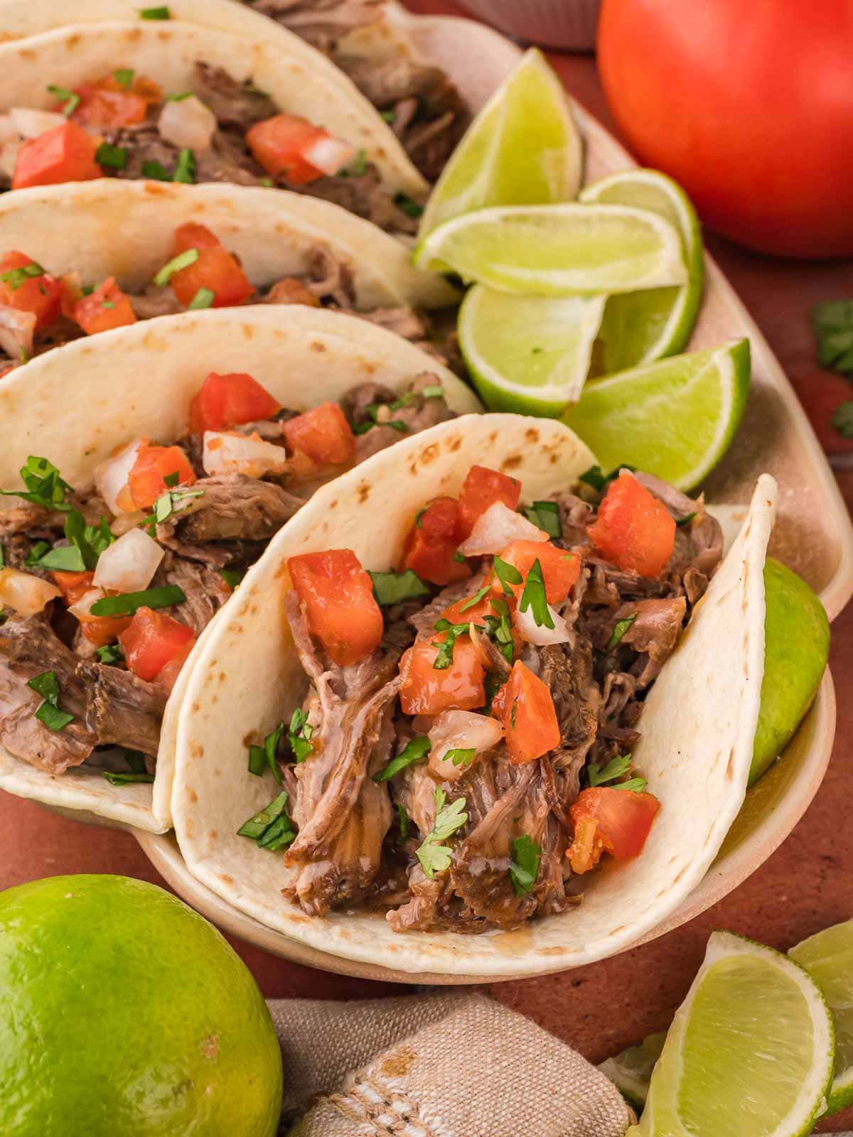 Plate of shredded beef tacos with diced tomatoes, onion, and cilantro, served with lime wedges.