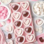 Assorted homemade filled chocolate hearts arranged on a pink tray with candy melts and sprinkles around them.