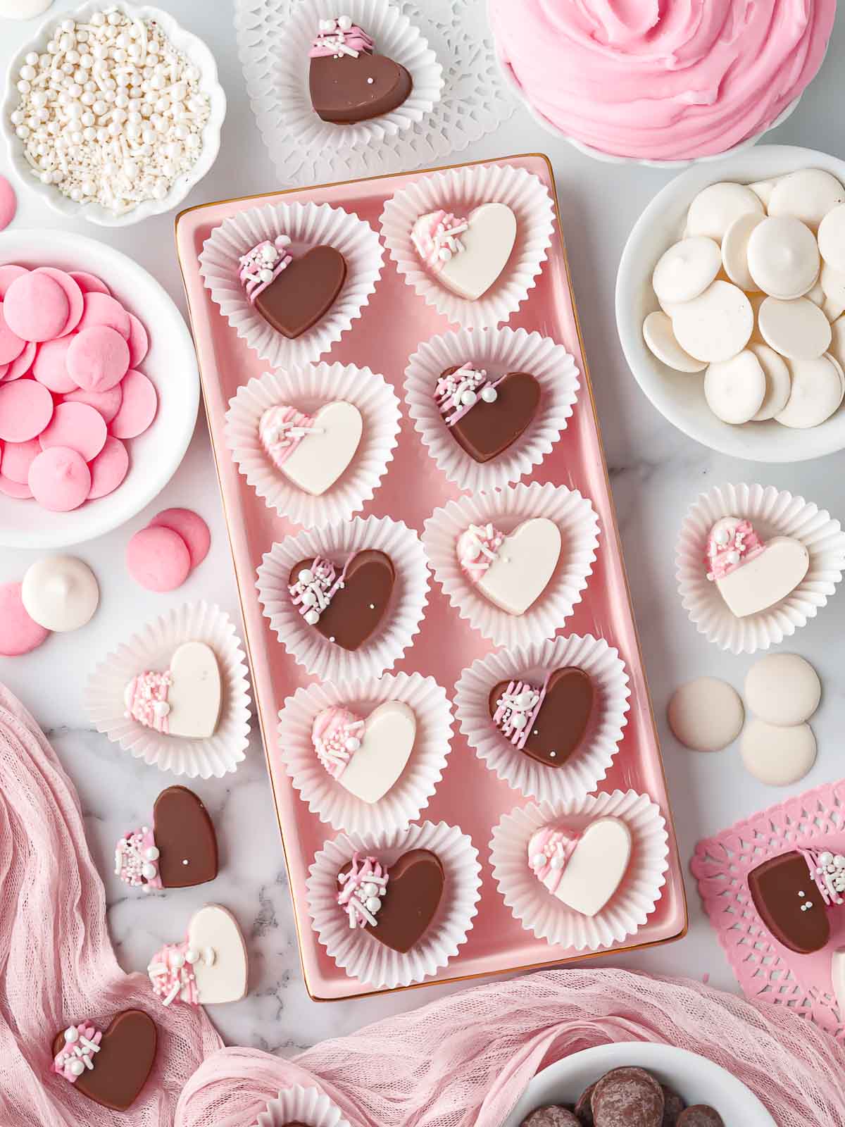 Assorted homemade filled chocolate hearts arranged on a pink tray with candy melts and sprinkles around them.