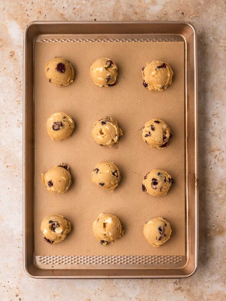 Scooped white chocolate cranberry cookie dough balls on a lined baking sheet.