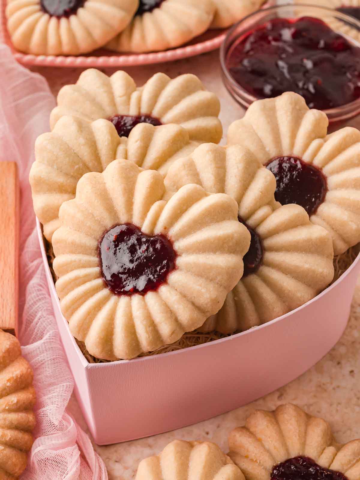 Heart jam cookies arranged in a pink heart-shaped box with a bowl of jam in the background. Close-up of a heart jam cookie showing th