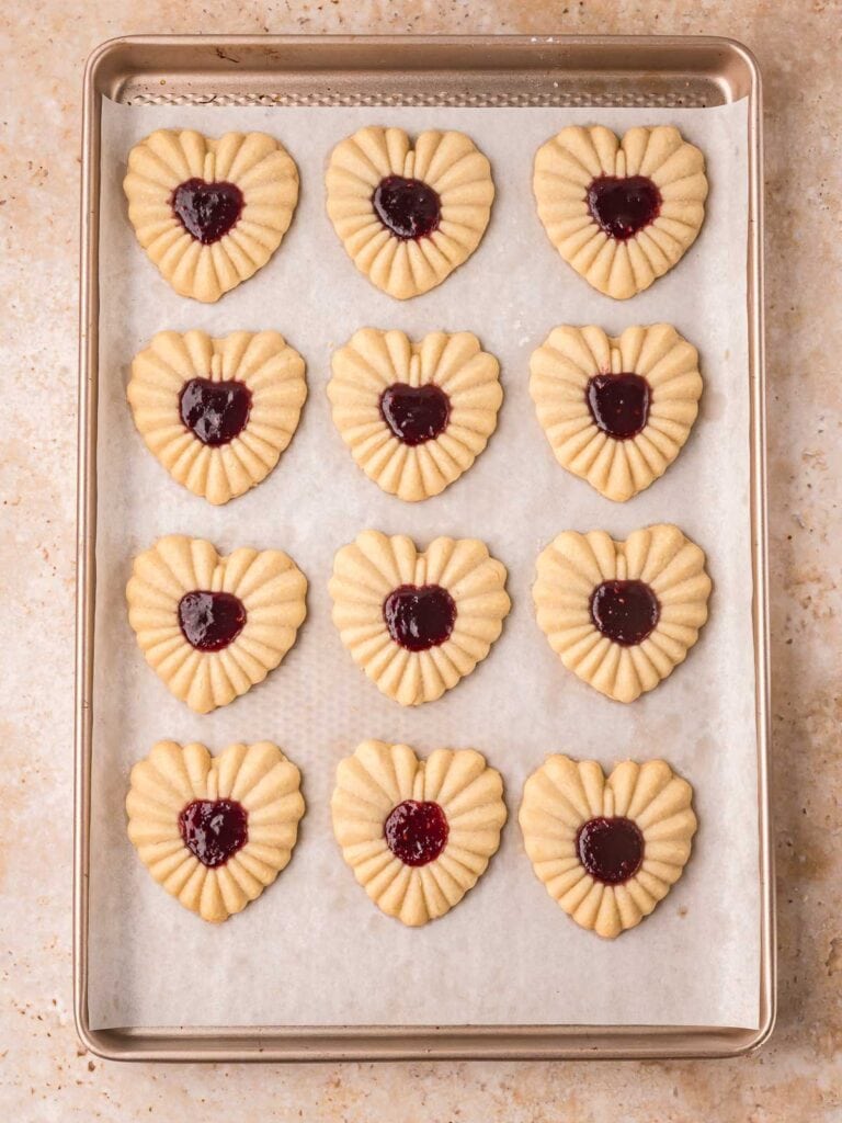 Heart-shaped stamped cookies arranged on a parchment-lined baking sheet before baking. Jam-filled heart cookies on a baking sheet, filled halfway through baking. Fully baked heart jam cookies with set jam centers cooling on the baking sheet.