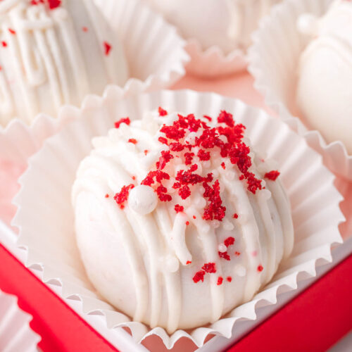 Red velvet truffle in a white paper liner inside a red gift box with white chocolate drizzle and red cake crumbs on top.
