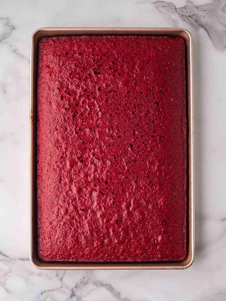 Baked red velvet cake cooling in a rectangular baking pan on a marble countertop.