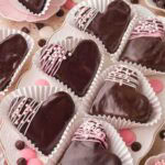 Chocolate-covered heart brownies arranged in white cupcake liners with Valentine sprinkles and pink accents.