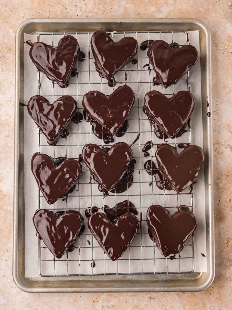 Chocolate-dipped heart brownies resting on a wire rack with excess chocolate dripping below.