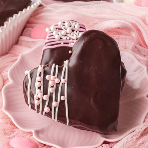 Close-up of a chocolate-dipped heart brownie with pink drizzle and white sprinkles on a pink plate Heart-shaped brownies coated in chocolate displayed on a pink background with Valentine candies.