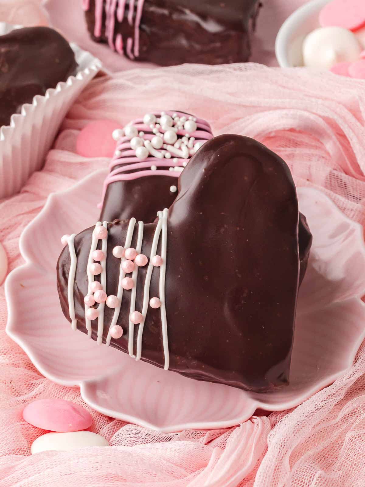 Close-up of a chocolate-dipped heart brownie with pink drizzle and white sprinkles on a pink plate Heart-shaped brownies coated in chocolate displayed on a pink background with Valentine candies.