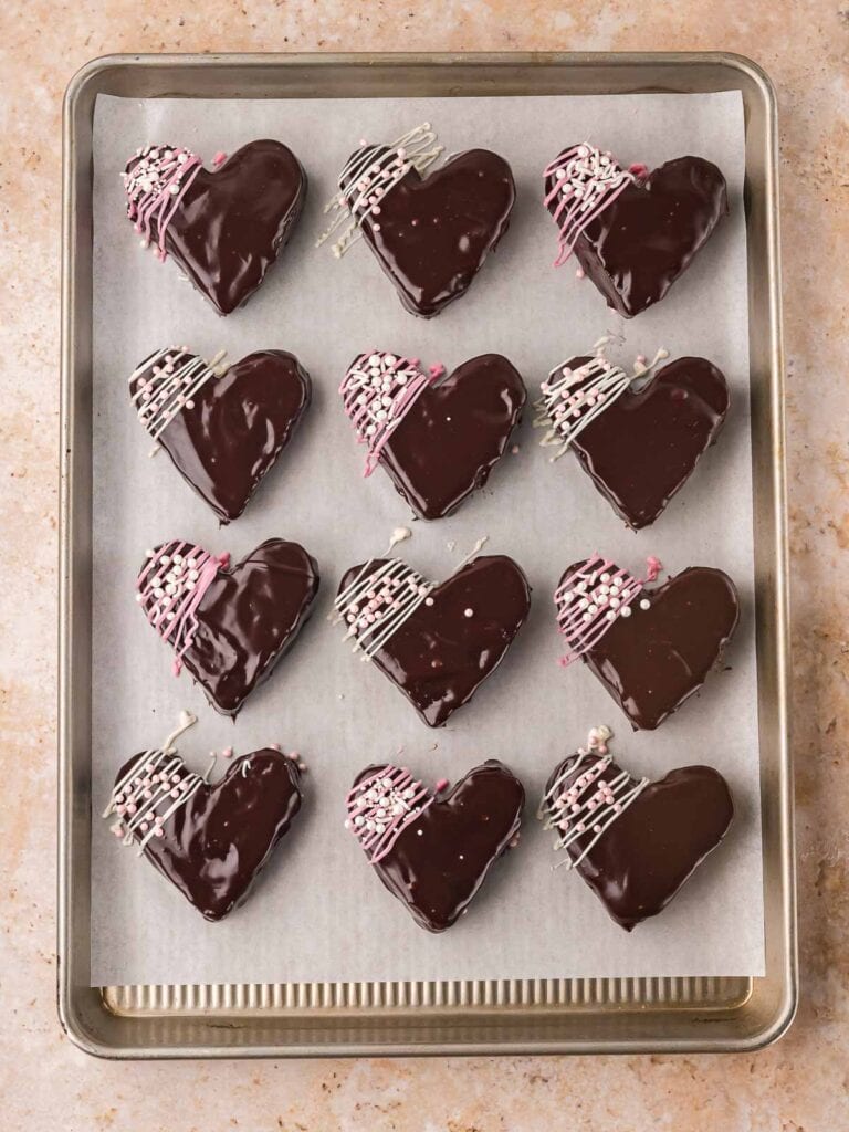 Chocolate-dipped heart brownies arranged on a parchment-lined baking sheet with pink and white drizzle.
