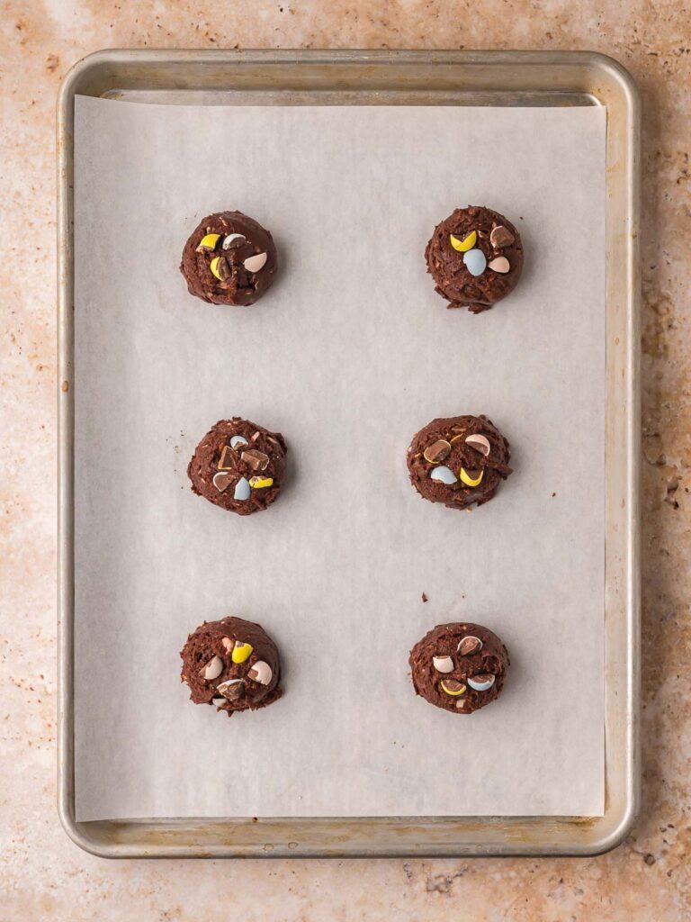 Scoops of Cadbury Egg brownie cookie dough placed on a parchment-lined baking sheet ready to bake.