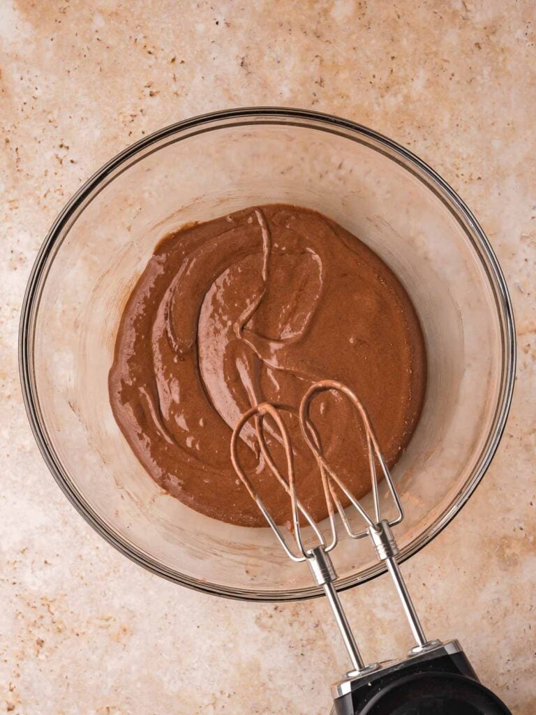 Chocolate brownie batter being mixed with a hand mixer until smooth and glossy.
