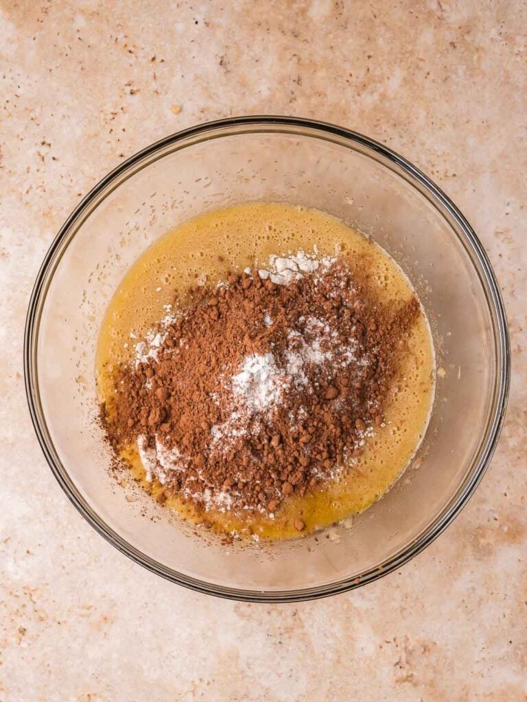 Cocoa powder and flour added to wet ingredients in a mixing bowl for brownie batter.