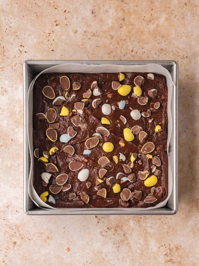 Brownie batter with Cadbury eggs spread into a parchment lined baking pan before baking.