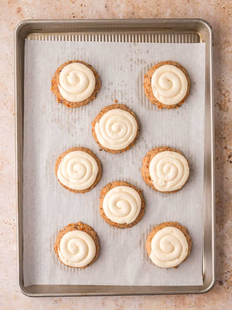 Baked carrot cake cookies topped with swirled cream cheese frosting on a baking sheet.