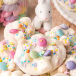 Easter candy clusters on a plate with pastel chocolate candies and sprinkles and a small bunny decoration in the background.
