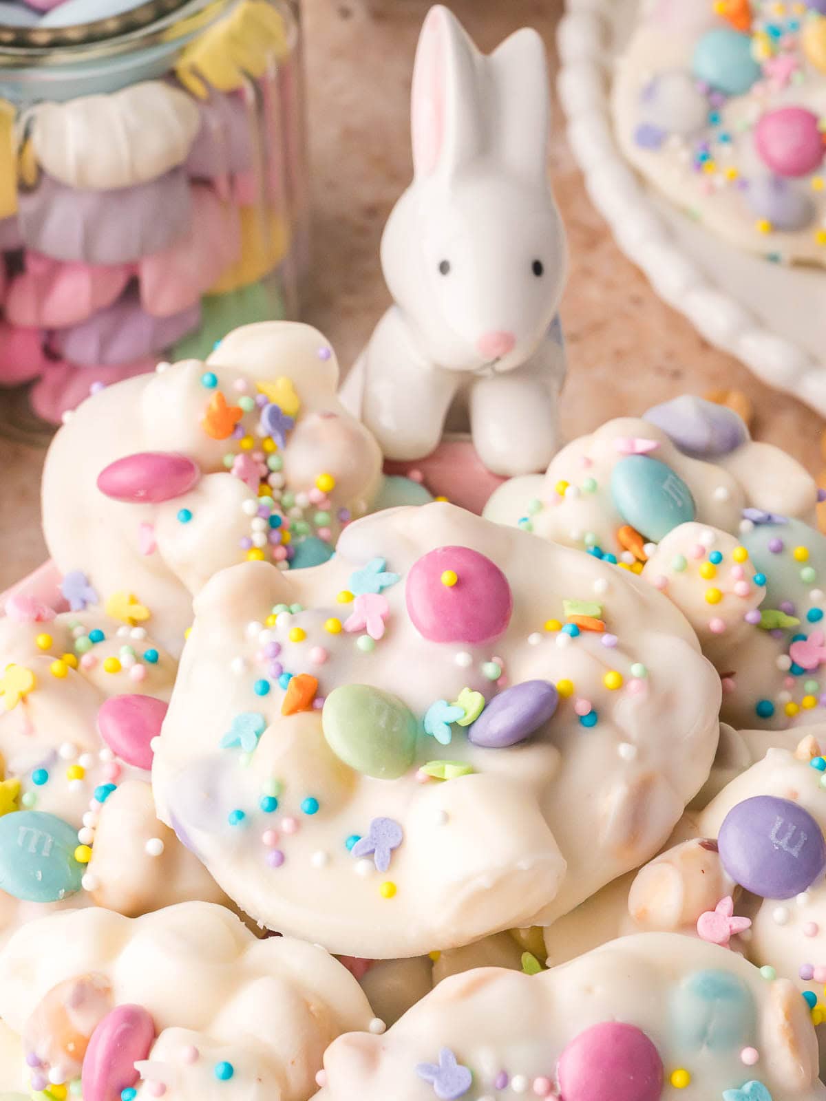 Easter candy clusters on a plate with pastel chocolate candies and sprinkles and a small bunny decoration in the background.