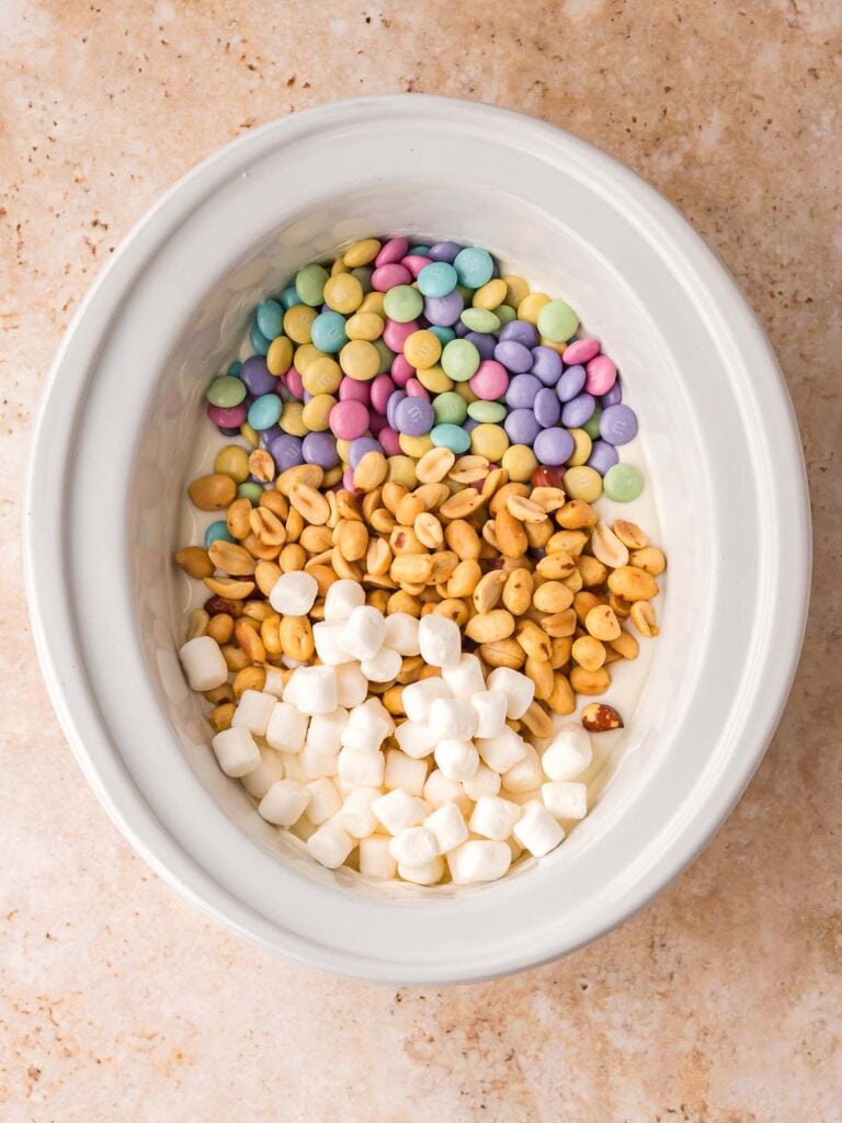 Slow cooker filled with white chocolate, peanuts, pastel M and M’s, and mini marshmallows before mixing.