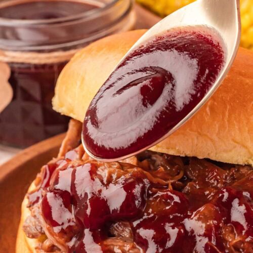 Spoon pouring thick homemade BBQ sauce over shredded beef sandwich.