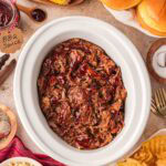 Slow cooker BBQ beef in crockpot surrounded by buns corn and sides.