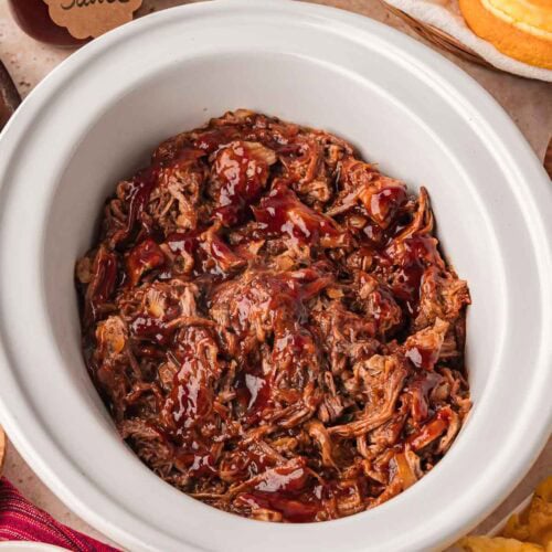 Top view of shredded BBQ beef in slow cooker with sandwich buns and sides.