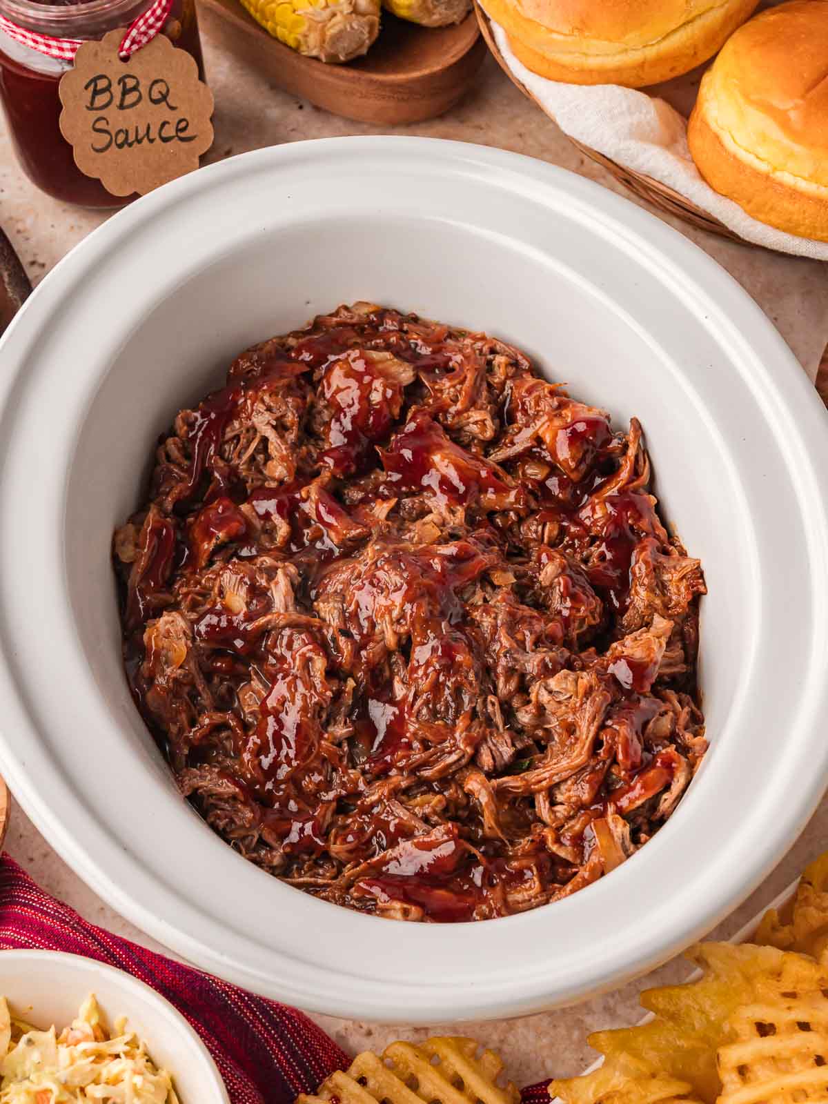 Top view of shredded BBQ beef in slow cooker with sandwich buns and sides.
