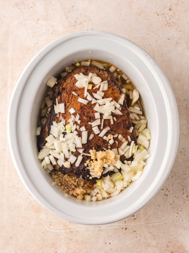 Seared beef placed in slow cooker with chopped onions and garlic.