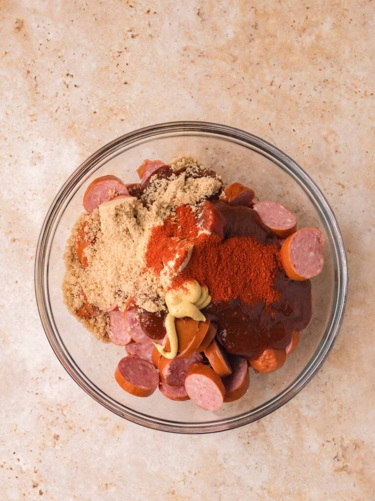 Smoked sausage pieces with BBQ sauce, brown sugar, and seasonings in a mixing bowl.