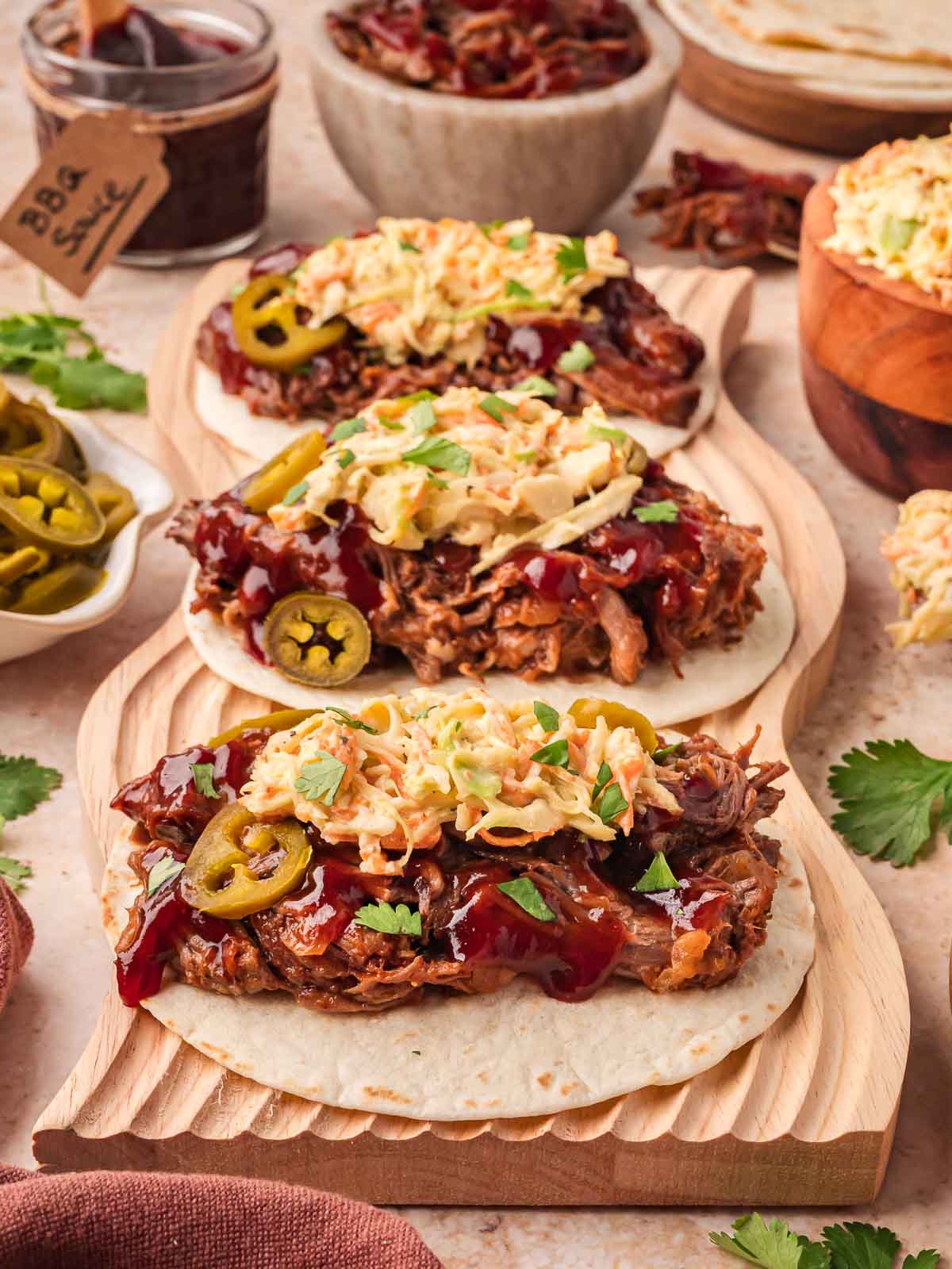 Three slow cooker BBQ beef tacos topped with coleslaw, jalapeños, and cilantro served with BBQ sauce and tortillas on the side.